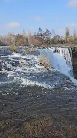 The video shows a wide waterfall with water cascading over a broad, flat edge into a churning pool below.  Mist rises from the base of the falls. The water above the falls is turbulent and dark. The surrounding area is a mix of bare trees and green shrubbery atop the cliffs. The sky is blue with a few scattered clouds. Buildings can be seen in the distance on the far side of the pool.