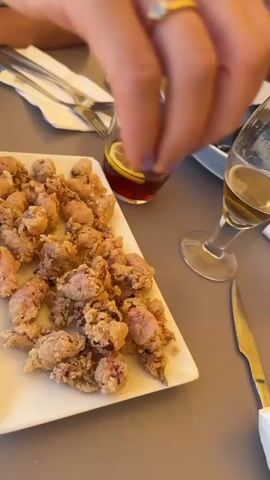 This is a close-up, over-the-shoulder shot of a table setting featuring a plate of fried calamari and drinks. The calamari, golden brown and lightly breaded, fills a rectangular white platter. In the background, several glasses are visible: one a wine glass with a pale liquid, another a short tumbler with a dark amber liquid and a lemon slice, and a third partially obscured wine glass with a similar pale liquid. A silver knife rests on the table to the right, and cutlery is arranged to the left. A bowl of dark mussels sits behind the calamari. The hand of a person is in the foreground, squeezing a wedge of lemon over the wine glass containing the pale liquid. The scene appears to be set outdoors, with ample natural light. The table surface is a muted gray, and the overall impression is a casual meal, possibly lunch or early dinner, at a restaurant or tapas bar. The text on the tumbler reads "VERMOUTH."