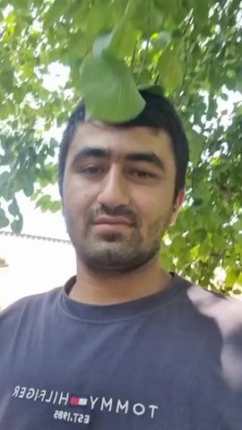 A man is in front of the camera outdoors. He has short dark hair, dark eyes, and a short beard. He is wearing a dark t-shirt with a logo on it that is difficult to read. He is looking directly at the camera. The background is out of focus, but it appears to be trees and some sort of building.