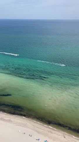 The video provides an aerial view of a beach and the ocean. The sand is light colored, and there are a few small figures scattered across it, possibly people relaxing. The water transitions from shallow, clear green near the shore to a deeper blue further out. There are two jet skis visible in the water, creating white wakes as they move. The sky is a pale blue with some wispy clouds. The overall scene depicts a sunny day at the beach with recreational activity.
