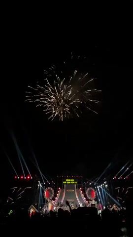 The image depicts a grand nighttime celebration in Măng Đen, Vietnam. Bright, golden fireworks explode high in the dark sky, casting a warm glow. Below the fireworks, a large, elaborate stage is brightly illuminated with numerous spotlights, several of which project strong beams upwards into the night. On the stage's central screen or backdrop, the Vietnamese text "MĂNG ĐEN KHÁT VỌNG VƯƠN XA" is clearly visible, likely indicating the event's location and theme ("Măng Đen - Aspiration to reach far"). In front of the stage, a crowd of people is silhouetted, suggesting a large audience gathered to watch the performance and the fireworks display. The scene conveys a festive and lively atmosphere.