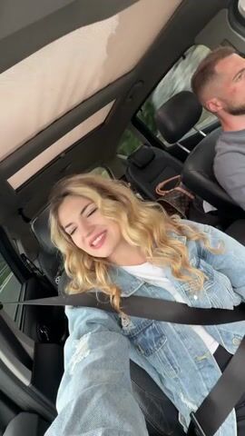 A woman is singing in the passenger seat of a car, in a selfie-style video. She has long, wavy blonde hair and is wearing a denim jacket over a white t-shirt. She has a seatbelt on. A man is driving the car. He has short brown hair and a beard and is wearing a gray shirt. The car has a sunroof and black leather seats. A brown purse is in the backseat.