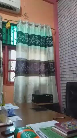 The video shows an office environment. In the foreground is a desk with various papers and books. To the left of the desk is a printer. A chair is positioned behind the desk. To the left of the desk, a window is visible with green bars and curtains. An air conditioner is mounted on the wall above the window. The wall to the right of the window is pink, and further to the right, the wall is covered with a brick-patterned wallpaper. A bulletin board with several documents is mounted on the brick-patterned wall. The video appears to be a static shot with no movement.
