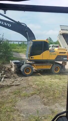 This video shows a yellow Volvo EW205D wheeled excavator operating in an outdoor industrial or construction setting. The excavator is actively digging and moving a pile of rubble and dirt with its bucket. In the background, another vehicle, possibly a dump truck with its bed raised, is visible. Trees and overgrown vegetation are present, suggesting an area that might be undergoing demolition or reclamation. A large, industrial-looking building with rusted elements is partially visible on the left side of the frame. In the distance, a bridge or overpass can be seen. The sky is bright and appears to be daytime with some clouds, indicating fair weather. The overall scene depicts a construction or demolition activity. The text "VOLVO" is clearly visible on the excavator's boom, and the model designation "EW205D" is present on the side of the machine. Other visible text includes "VOLVO" on the cab and what appears to be a company logo or name next to it.