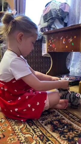 A young girl with blonde hair pulled into a ponytail is sitting on a patterned rug. She is wearing a white t-shirt and a red dress with white hearts. The girl is focused on a stuffed animal, possibly a cat, which is grey and black striped. She is holding a pink item, possibly a toy or a utensil, and interacting with the stuffed animal. To her right, there is a wooden piece of furniture adorned with colorful stickers. In the background, there is a purple curtain and a pile of blankets. The environment appears to be indoors, likely a child's room or living area.