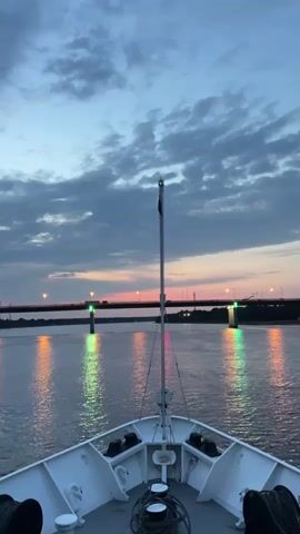 The video shows the bow of a white boat moving forward on a body of water. The sky above is cloudy with a sunset visible in the distance. There is a bridge crossing the water ahead, with lights on it and on buoys in the water. The boat appears to be moving towards the bridge.