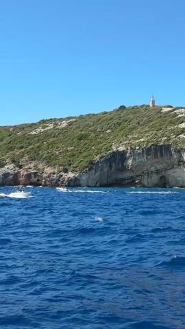 The video shows a boat moving on the blue ocean towards a rocky coastline. The coastline is covered with green vegetation, and there are caves carved into the cliff face. On the highest point of the hill, a white lighthouse can be seen. The sky is clear and blue, indicating a sunny day. Two boats are visible in the distance, moving on the water closer to the shore.