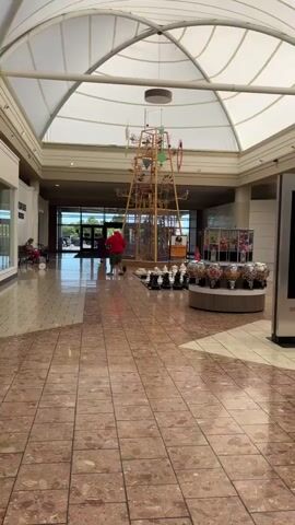A man in a red shirt and black shorts walks away from the camera in a brightly lit indoor shopping mall. He is walking past a display of gumball machines and a tall, elaborate kinetic sculpture. To the right of the sculpture is a digital information kiosk. Further to the right is a Kay Jewelers store. The floor is tiled with large, square, reddish-brown tiles. The ceiling is a white, vaulted canopy.
