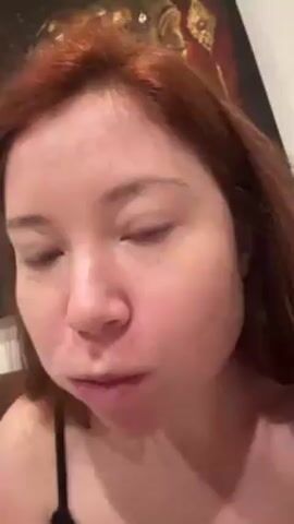A woman with red hair eats a hamburger in a close-up shot. The woman is wearing a black tank top, and there is a painting on the wall behind her. She takes a bite of the hamburger, chews, and smiles.