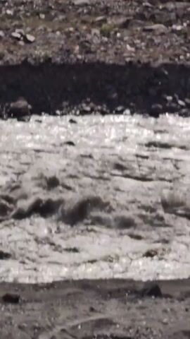A video shows a muddy river flowing through a rocky, mountainous landscape. The river is churning with white water in some areas, suggesting a fast current. The banks of the river are lined with rocks and dirt, and in the background, there are steep, green mountains under a bright blue sky with some scattered clouds. A road or path is visible on the riverbank in the foreground.