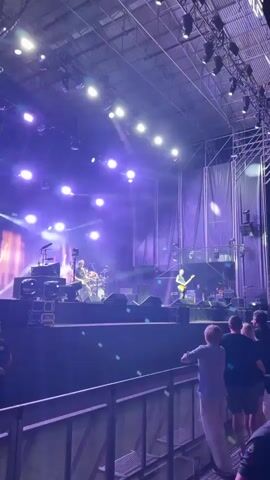 The video captures a live concert performance. A band with at least three musicians, a drummer and two guitarists, are visible on a dimly lit stage bathed in purple and white spotlights. They are performing on a raised platform with various musical instruments and audio equipment. In the foreground, a crowd of concertgoers stands behind a metal barrier, some looking towards the stage and others interacting with each other. The overall atmosphere suggests an energetic and engaging musical event.
