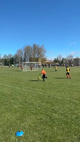 A children's soccer game is captured on a bright, sunny day on a green outdoor field in Ragana, Latvia. Several young players, mostly in orange, yellow, and black jerseys, are actively involved in the match. The camera is positioned low to the ground, with a blue cone visible in the immediate foreground.

In the mid-ground, a player in an orange jersey kicks the soccer ball towards the goal. The goalkeeper, wearing a yellow jersey, attempts to block it but the ball goes into the net, scoring a goal. Other players are scattered across the field, with some near the goal and others further back.

In the background, more people, including adults and children, are visible observing the game. Trees line the perimeter of the field, and a building can be seen in the distance under a clear blue sky. The atmosphere appears lively, with cheers and shouts audible in the background as the goal is scored.