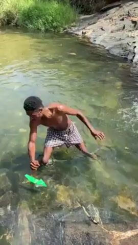 A dark-skinned young man in a shallow, clear stream of water kneels on a rock and splashes water. A green plastic object floats nearby. He sits down in the stream. The camera pans to the left to show water flowing over rocks.