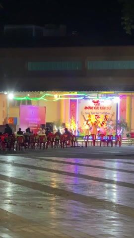 A long shot depicts an outdoor stage during a nighttime event in Vietnam. On the stage, a banner reads "ĐỜN CA TÀI TỬ" with additional Vietnamese text, indicating a musical performance. Colorful decorations and balloons frame the stage. A woman in a light-colored dress is standing in front of the microphone, possibly performing or about to perform.

Several rows of red plastic chairs and tables are arranged in front of the stage, where a group of people are seated, appearing to be an audience. They are mainly silhouettes due to the backlighting from the stage. A security guard can be seen standing near the stage.

The ground is paved with large, rectangular stones, and there is some reflective moisture on the surface, likely from a recent rain. The light from the stage and surrounding area is reflecting off the wet pavement, creating a shimmering effect.

The background features a low building with windows, some of which are lit. Some branches of trees are visible at the top of the frame on either side. A person in a hardhat and a light-colored uniform rides a scooter in front of the camera and past the stage, blurring the camera's point of view briefly.