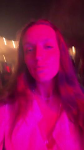 A young woman is seen in a close-up video, moving and singing along to music. The lighting changes between red, blue, and purple, creating a dynamic atmosphere. She's wearing a white shirt and a necklace. There is a blurry figure of a man behind her in the initial frames. In the background, indistinct lights suggest she is outdoors, possibly at a festival or party.