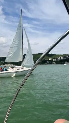 The video shows a white sailboat with two white sails on a body of water. The sailboat has a few people on board. The water is green and there are hills in the background with some buildings on them. The sky is blue with some white clouds. The camera appears to be on another boat, looking towards the sailboat.