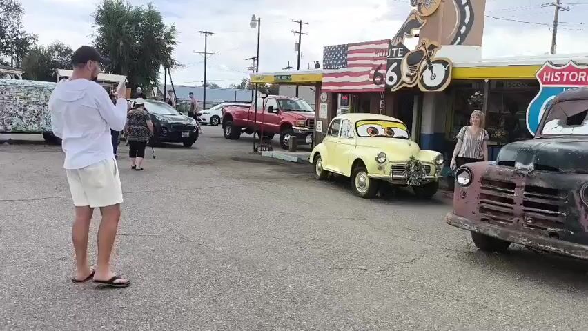 This video shows a street scene at what appears to be a roadside attraction or gift shop on Historic Route 66. In the foreground, a man wearing a white shirt and shorts is holding up a phone, seemingly taking a photo of the scene. To his right, a dark-colored truck with cartoon-like eyes and a weathered appearance is parked. Further back, a small, pale yellow car, also with cartoon eyes, is positioned in front of a building with a yellow awning. The building has signs for "Route 66" and "Historic." There are other vehicles in the background, including a red pickup truck and a black SUV. A woman with short, gray hair is standing near the yellow car, looking towards the camera. To the right of the dark truck, a purple station wagon with cartoon eyes is parked. The ground is paved, and there are trees and utility poles visible in the background. The overall impression is a display of vintage vehicles, possibly themed around the movie "Cars," situated along a historic highway.