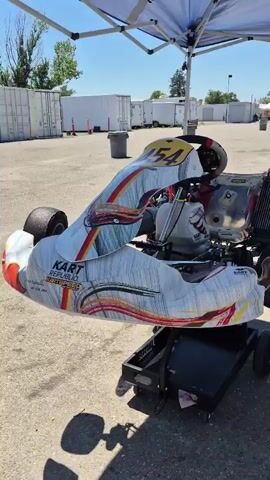 The video shows a go-kart on a kart stand outdoors. The go-kart is white with colorful racing graphics. The front of the go-kart has the number 54 on it, and the rear has the number 454. The go-kart has a racing seat, steering wheel, engine, and radiator. The background shows shipping containers, a tent, and some trees. The video appears to be taken on a sunny day.
