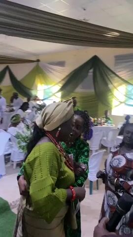 The video shows a group of people at what appears to be a wedding reception. The room is decorated with green and white fabric draping from the ceiling. Many people are wearing green and white clothing, some with traditional head wraps. Two women in the center are talking and smiling while holding a piece of paper, potentially a program or photograph. A man approaches them and hands one of the women what looks like money. The atmosphere seems celebratory.