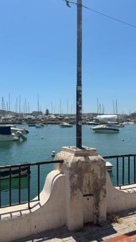 The video pans across a harbor filled with numerous boats and yachts under a clear blue sky. In the foreground, a low wall with a black metal railing is visible, with a lamppost on the right side. The water is a calm, turquoise color. Several boats are moored in the harbor, including speedboats and sailboats. In the background, buildings line the shore, and more boats are docked further in the distance. A palm tree is present in the lower left corner of the frame in the last few seconds. The scene appears to be a sunny day in a coastal town or city.