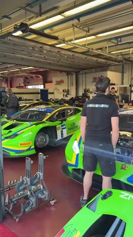The video shows a pit garage with several bright green Lamborghini race cars. The cars are the primary focus, with their hoods open and some appearing to be undergoing maintenance. Several individuals, dressed in casual attire and some in team shirts, are present in the garage. Some are looking at the cars, while others appear to be working on them. The environment is a typical race pit garage, with overhead lighting, metal structures, and garage doors visible in the background. The cars are decorated with racing decals and sponsors' logos.