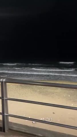 A video shot at night shows a sandy beach with waves breaking. A metal railing runs along the edge of the beach, separating it from a brick walkway. Streetlights illuminate the walkway, casting long shadows. Several people are seen walking along the walkway, and one person is sitting on a bench. In the distance, buildings and lights are visible. The camera pans left, revealing more of the walkway and the ocean.