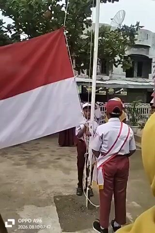 The video shows a flag-raising ceremony outdoors. Two young students, dressed in school uniforms with red and white accents, are at the base of a flagpole. One student on the left holds the Indonesian flag, which is red and white, partially unfurled. The other student on the right, wearing a sash over their uniform, appears to be operating the flagpole mechanism. Other students in similar uniforms are visible in the background, some standing at attention. The environment includes trees, a building with a sign, and other structures. The ground is paved. The timestamp overlay indicates the video was recorded on August 10, 2026.