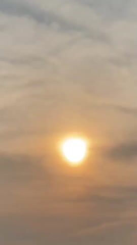 A short video shows the sun partially obscured by clouds at sunset. The sun is bright and appears as a golden orb in the sky. The clouds are wispy and scattered, with a warm, orange glow. The lower portion of the video shows a silhouette of trees and vegetation against the sky. The overall mood is peaceful and serene.