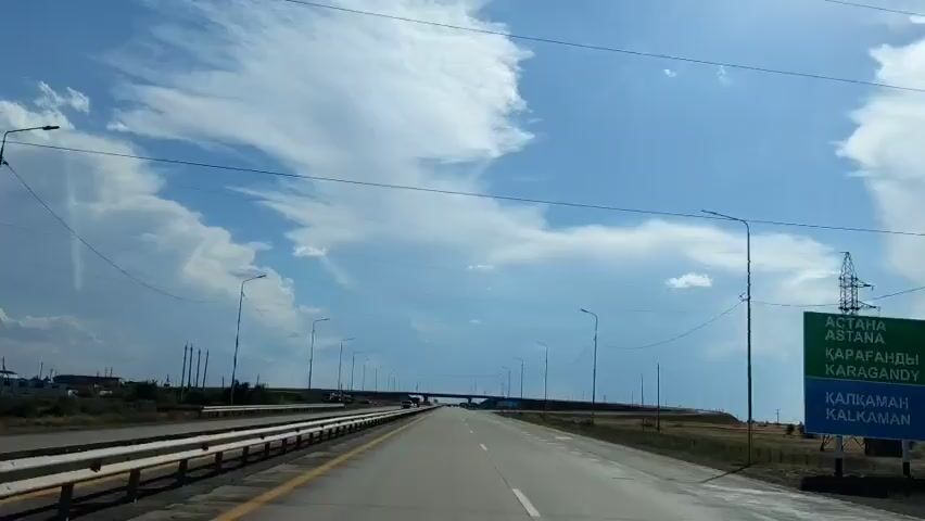 The video shows a view from a moving vehicle on a highway under a cloudy blue sky. The road is paved and has two lanes in each direction, separated by a yellow line. There are guardrails on the left side of the road and numerous streetlights lining the highway. A bridge crosses over the highway in the distance. A few cars and trucks can be seen on the road. The landscape on either side of the highway appears to be flat and grassy, with some scattered trees and power lines. The sun is shining, casting shadows on the road.