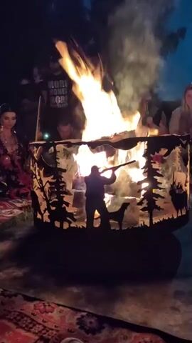 A fire pit with a metal screen depicting a forest scene with a hunter and a dog is burning brightly at night. Several people are gathered around the fire pit, some of whom are visible in the background. The fire pit is placed on a patterned rug. A woman in a patterned dress is seated to the left of the fire pit. The overall atmosphere appears to be a social gathering around a bonfire.