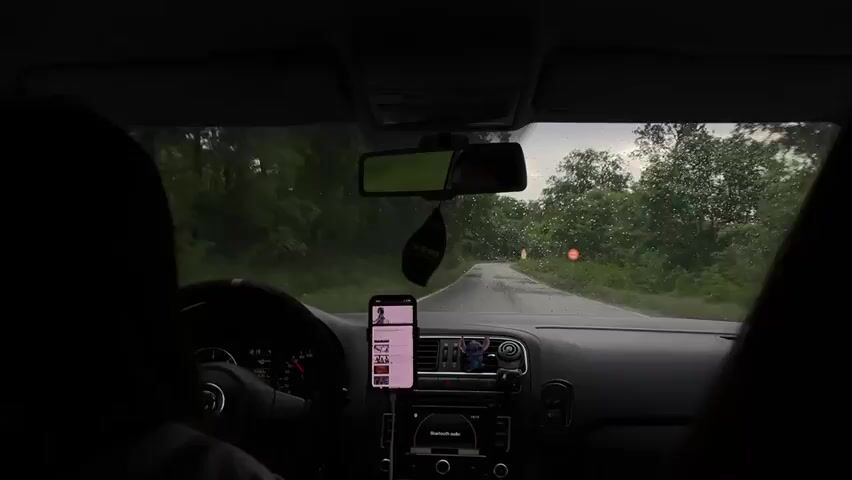 A car drives down a road on a rainy day. The view is from the back seat, looking out the front windshield. A cell phone is mounted on the dashboard, displaying a playlist. The car is black, with a leather steering wheel. An air freshener hangs from the rearview mirror.  The road is narrow and winding, surrounded by trees. The sky is overcast.