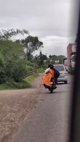 A person is riding a motorcycle on a road with another person on the back. They are wearing an orange dress. There are trees and greenery on the side of the road, and a cloudy sky above. In the background, there are cars and trucks on the road.