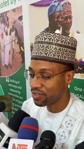 A man with glasses and a decorated hat speaks into a cluster of microphones, including one labeled NTA. He wears a white shirt with geometric designs around the collar. Behind him is a poster with text and images, including portraits of two men in traditional clothing. One section of the poster mentions "Desilting of waterways". The man appears to be giving an interview.
