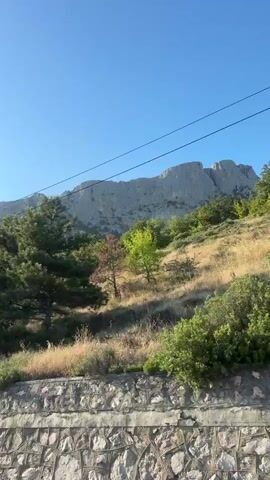 This video shows a view of a mountainside covered in trees, with a clear blue sky above. The camera appears to be moving, possibly from a vehicle, as the scenery shifts. There are power lines visible in the upper portion of the frames, and a stone wall or barrier is seen in the foreground of the earlier frames. The sun is shining, creating a bright and clear atmosphere. The vegetation consists of various shades of green, with some trees appearing taller and more mature than others. The mountains in the background are rugged and rocky, with their peaks extending upwards towards the sky. The video offers a glimpse of a natural, mountainous landscape bathed in sunlight.