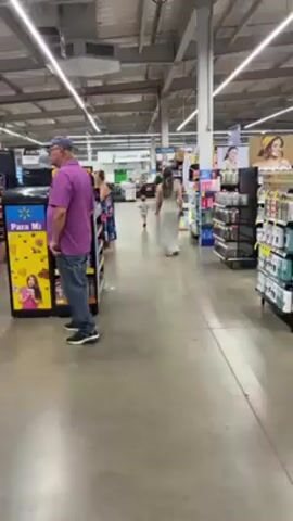 The video shows the interior of a Walmart store in Honduras. Several customers can be seen browsing the aisles, including a man in a purple shirt and jeans, a woman in a floral dress, and a woman in a white dress with a child. The aisles are stocked with various products, including snacks, beverages, and personal care items. The store has a typical warehouse-style ceiling with exposed ductwork and fluorescent lighting.