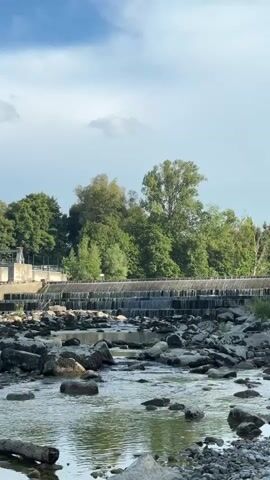 A wide shot shows a rocky riverbed with a dam in the background. The river flows gently over the rocks. The dam has a series of cascading waterfalls. Behind the dam, a line of lush green trees forms a backdrop. The sky above is partly cloudy with patches of blue. The scene is calm and natural.