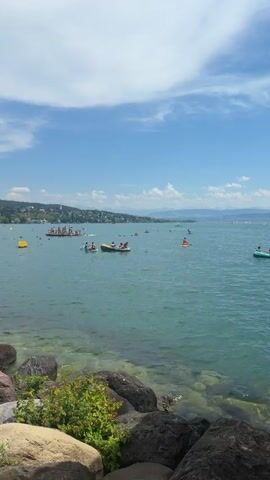 This video captures a lively scene at a lake on a sunny day. In the foreground, large rocks line the shore, with some green foliage growing between them. The water is a clear turquoise, reflecting the bright blue sky dotted with white clouds. The middle ground is filled with activity on the water. Several small boats, including what appear to be kayaks or canoes, are visible. Some have multiple occupants, suggesting recreational use. There are also yellow buoys scattered across the lake, possibly marking swimming areas or channels. In the background, a town or city stretches along the shoreline, with buildings nestled amongst trees. A long, narrow boat with many people rowing in unison is a prominent feature. Numerous individuals are also swimming or wading in the water closer to the shore. The overall atmosphere is one of a bustling summer day at a popular recreational spot.