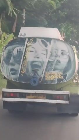 A rear view of a tanker truck is shown in motion. The truck is green and white, with a large, oval-shaped tank painted with three portraits of women. The women are wearing sunglasses and have their mouths open, as if singing or shouting. One portrait in the center is of a woman with her mouth wide open in a scream or laugh, and her eyes are closed. There is a blue valve on the side of the tank. A license plate is visible on the back of the truck. The truck is on a road, with trees and foliage visible in the background. The video is blurry due to the motion of the truck.