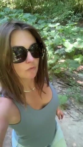 A woman with brown hair and sunglasses is walking in a wooded area. She is wearing a light blue tank top and a denim skirt. The camera is positioned at a low angle, capturing her from the waist up. The background consists of trees and foliage. The lighting suggests it is daytime.