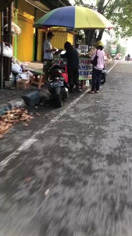 The video shows a street scene with several people and objects. In the foreground, a red motorcycle is parked. To the right of the motorcycle, a food stall with a colorful umbrella is set up. Three people are interacting at the food stall. One person, wearing a dark jacket and a helmet, is standing behind the stall, possibly preparing food. Another person, wearing a light-colored shirt and a hat, is standing in front of the stall, seemingly interacting with the vendor. A third person, wearing a pink jacket and a helmet, is also standing near the stall. The environment appears to be outdoors, with a paved road and a building in the background. The lighting suggests it might be daytime, possibly early morning or late afternoon. There are also some items on the ground near the food stall, and other motorcycles visible in the distance.