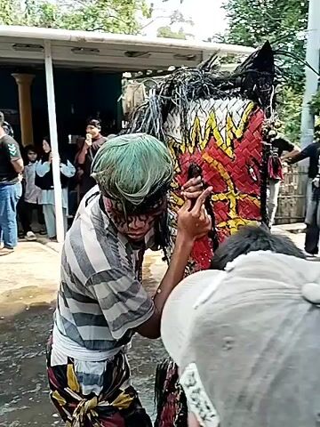 The video depicts a cultural performance or ritual involving a large, horse-like effigy. The effigy is brightly colored, with red, yellow, and black accents, and is adorned with dark, flowing material resembling a mane and tail. A young person, possibly a performer, is partially visible within the effigy, with their face and hair painted. They are adjusting or holding parts of the effigy. Other people are gathered around, observing the scene. Some are standing on the side, while others are closer to the effigy. The environment appears to be an outdoor setting, possibly a village or community gathering, with houses and trees visible in the background. The ground is wet, suggesting recent rain or water being used as part of the ritual. The overall atmosphere seems to be one of active participation and observation of a traditional event.