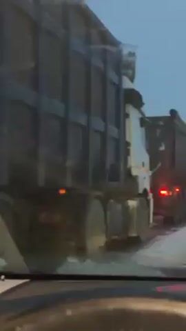 The video shows a driver's perspective from inside a car stuck in a line of large trucks on a road that appears to have snow or ice on it. The trucks are a mix of dump trucks and semi-trailers. The sky is overcast and the overall light suggests it is either early morning or late afternoon.  The camera is mostly stationary, and the focus is on the trucks in front of the car.
