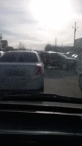 The video shows a series of views from inside a car. Initially, the view is through the windshield, looking out at a parking lot with several cars and utility poles visible in the distance. The scene then cuts to an exterior shot of a white mini-van with the driver's side door open. The camera angle shifts to capture the reflection in the side mirror, revealing a person in a brown jacket holding a phone. The video concludes with a view through the windshield again, showing a street lined with parked cars and overhead utility lines against a cloudy sky.