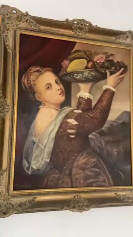 A painting is displayed in a gilded frame. The painting depicts a woman in a Renaissance-style dress holding a bowl of fruit. The woman has fair skin, blonde hair pulled back in an ornate hairstyle, and wears a pearl necklace. She is looking towards the viewer with a neutral expression. Her dress is dark brown with puffed sleeves and lace trim at the collar and cuffs. A light blue sash is draped over her left shoulder. The bowl of fruit she holds contains various fruits including grapes, pears, and flowers. Behind her, a dark red curtain is visible on the left, and a landscape with mountains is depicted on the right. The painting is signed in the lower left corner with illegible text and a date that appears to read "1974". The video pans slowly across the painting, first from left to right, then from right to left, showing the entire artwork and its frame. The painting is hanging on a white wall.