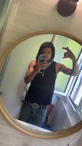 The video shows a young man with long, dark hair taking a selfie in a circular mirror. He is wearing a black tank top, blue jeans, and a studded belt. He is holding a smartphone in his right hand and making a hand gesture with his left. The background appears to be a bathroom with tiled walls and a window. The lighting is natural and the overall scene is casual.