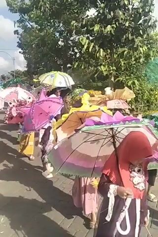 A group of people are walking in a parade outdoors. They are carrying and wearing a variety of colorful, decorative umbrellas. Some of the umbrellas are adorned with flowers, tinsel, and other embellishments. The people appear to be dressed in festive attire, and some are wearing hijabs. The ground is paved, and there are trees and foliage in the background. The overall scene suggests a celebration or cultural event.