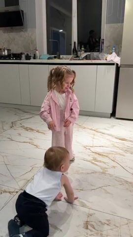 A little girl in a pink outfit dances in a kitchen with marble floors. She has her hair in pigtails and looks down as she dances, smiling. A baby lies on the floor behind her. There are white cabinets in the background with various items on top. The girl briefly looks away from the camera to her left, then back.