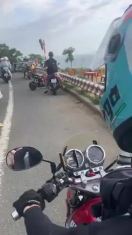 The video shows a red motorcycle stopped along a road overlooking the ocean. Other motorcycles and people are visible along the road to the left.  Orange plastic tables and chairs are set up near the roadside barrier. The camera is pointed toward the ocean, with land visible in the distance. The sky is partly cloudy.