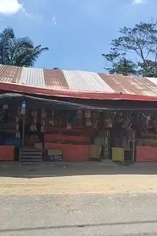 The video shows a view from a moving vehicle driving past a roadside shop. The shop has a corrugated metal roof, red and yellow boxes stacked up, and various items hanging from the roof. People are seen standing and moving around in front of the shop. The surroundings include trees, including palm trees, and other vegetation. The sky is blue with scattered clouds.