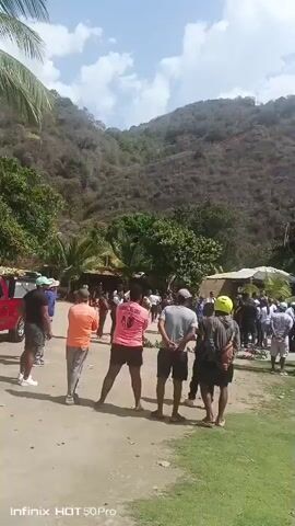 The video shows a group of people gathered at what appears to be a tropical resort or outdoor venue. The setting includes a grassy area, a mountain in the background, palm trees, and small, thatched-roof structures.  There is also a sign written in Spanish that says “Bienvenidos”. Some people are standing around, while others seem to be taking pictures.  In the distance, there are small, circular swimming pools. The weather appears sunny and partly cloudy.